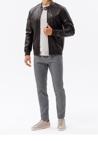 Muž v černé kožené bundě, bílém tričku, šedých úzkých kalhotách a béžových teniskách stojící na bílém pozadí.