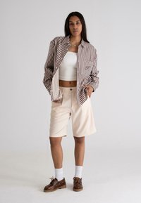 Stribet brun og hvid oversized skjorte med en lomme, hvid crop top, beige shorts, hvide sokker og brune snørebåndssko.