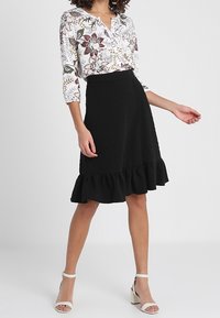 Bluse mit floralem Muster in Weiß und drei Viertelärmeln, kombiniert mit einem schwarzen, strukturierten knielangen Rock mit Rüschensaum und Riemchen-Pumps.