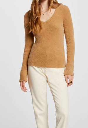 Femme portant un pull beige doux à manches longues avec col en V et un pantalon écru sur un fond uni.