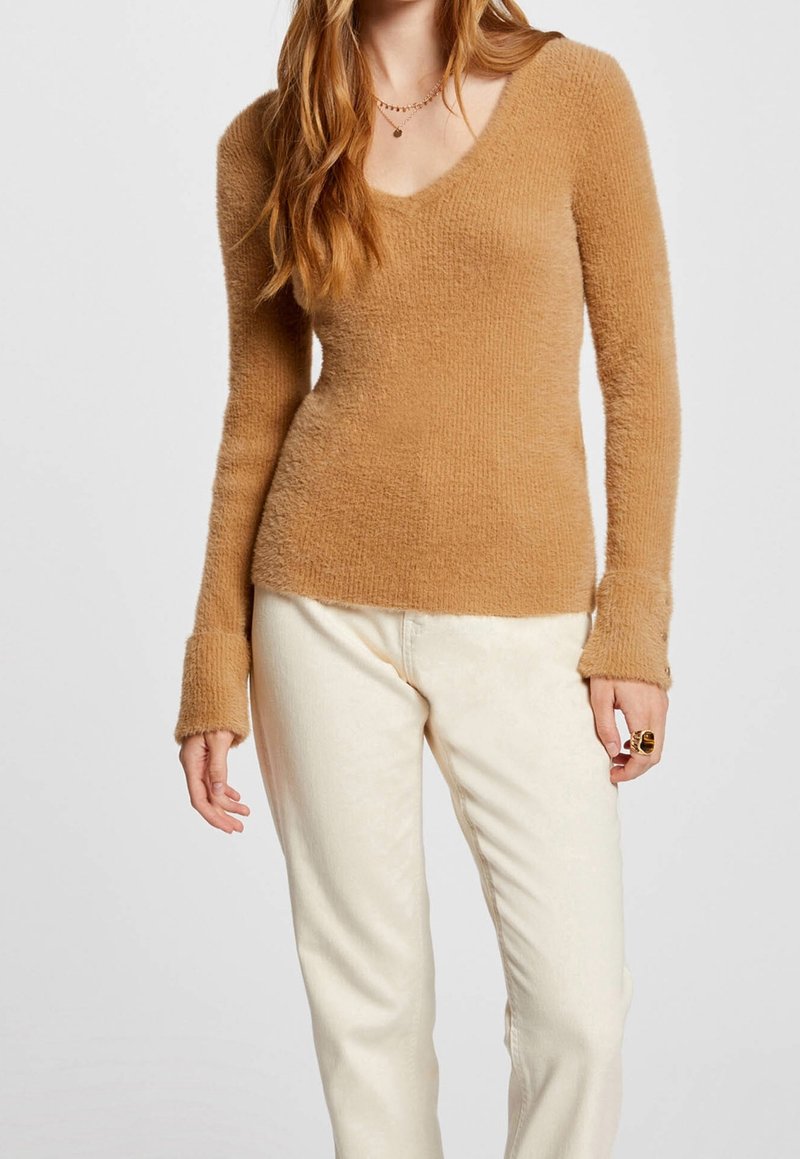 Femme portant un pull beige doux à manches longues avec col en V et un pantalon écru sur un fond uni.