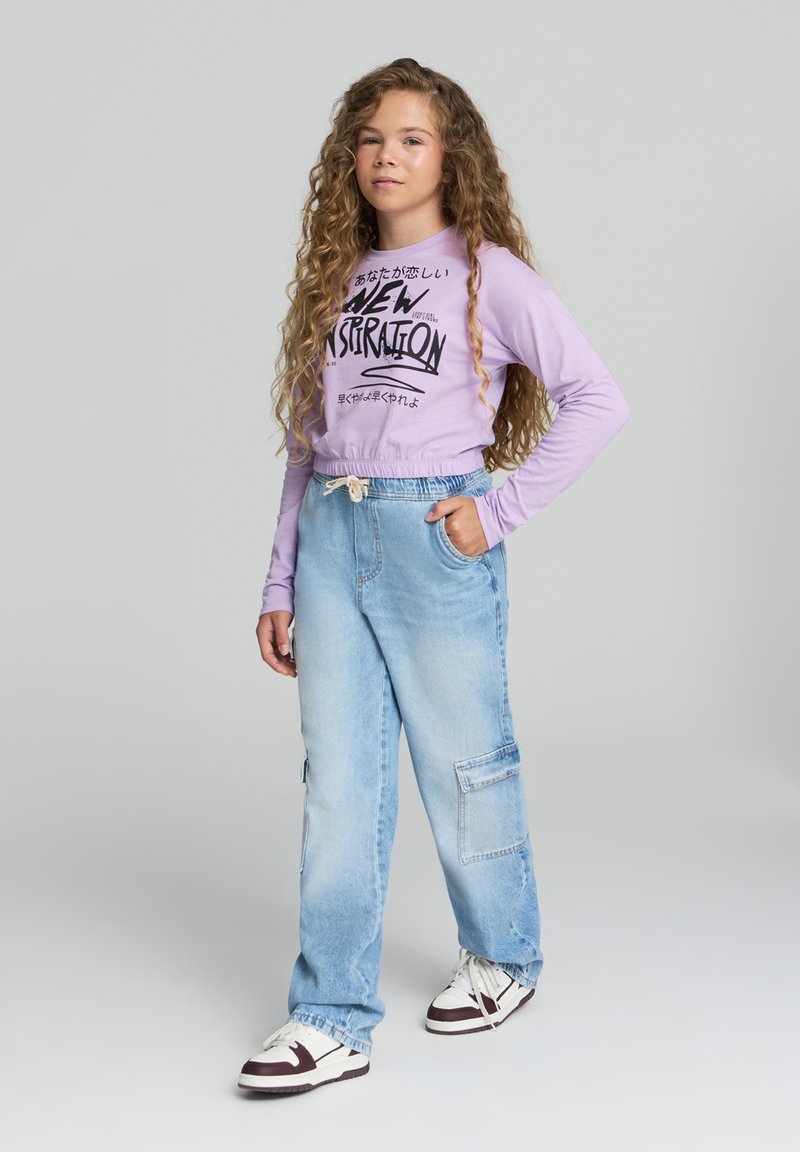 Liliowa, krótka koszula z długim rękawem z czarnym napisem graficznym, w zestawieniu z luźnymi, jasnoniebieskimi spodniami cargo dżinsowymi. Białe i ciemnobrązowe trampki.