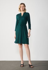 Teal mekko napitus edessä, pitkät hihat, säädettävä vyötärö ja polvipituinen hame. Yhdistetty mustiin, solmittuihin sandaaleihin.