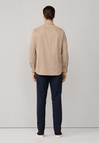 Beige overhemd met lange mouwen en een kraag, gemaakt van textuurstof, gecombineerd met donkerblauwe broek en witte sneakers, gezien vanuit de achterkant.