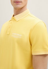 Gul polo-tröja med en strukturerad krage, tre knappar och vit logotryck på bröstet. Har kontrasterande ränder på ärmarna.