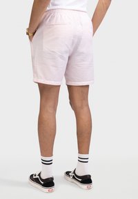 Άντρας που φοράει ροζ υφασμένα σορτς, λευκές κάλτσες με μαύρες ρίγες και μαύρα αθλητικά παπούτσια, εμφανιζόμενος από πίσω με τα χέρια στις τσέπες.