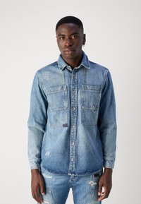 Camisa de mezclilla en azul claro con cierre de botones, dos bolsillos en el pecho y detalles desgastados en las mangas y el dobladillo. Mangas largas, cuello estándar.