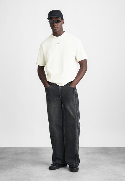 Tricou oversized de culoare bej deschis, blugi negri cu croială largă și detalii cusute pe lateral, șapcă neagră și ochelari de soare închiși la culoare. Încălțăminte de culoare neagră.