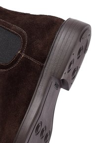 Bruine nubuck schoen met zwarte elastische zijkant en een donkere rubberen zool met cirkelvormige grippatronen.