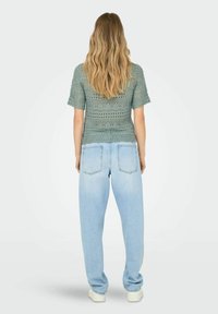 Top corto de punto verde con mangas cortas, que presenta un patrón de encaje y un dobladillo en forma de olas, combinado con unos jeans de cintura alta en azul claro.