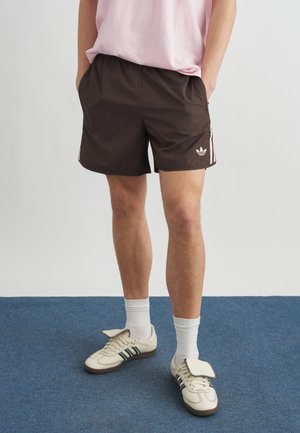 Person iført mørkebrune Adidas-shorts med pink striber, lyserød skjorte, hvide sokker og hvide Adidas-sneakers med sorte striber på blåt tæppe.