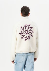 White fleece jacket with a high collar. Back features a purple embroidered sun and eye design. Relaxed fit with elastic hem and cuffs.