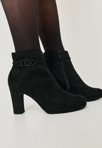 Botas ankle em camurça preta com um salto alto, apresentando um fecho de correr e um detalhe em corrente decorativa perto do topo. Textura suave, design moderno.