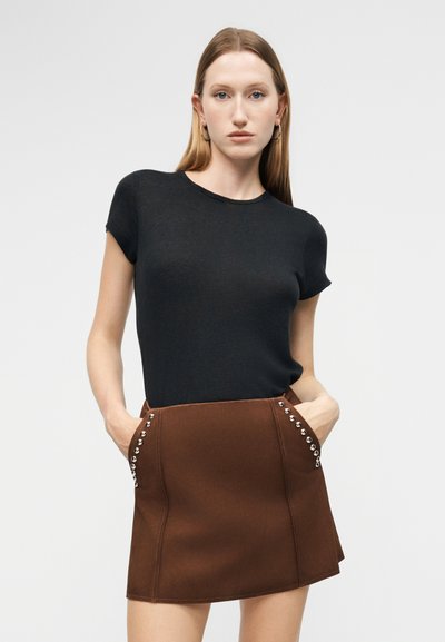 Νεαρή γυναίκα με μακριά ξανθά μαλλιά, φορώντας μαύρο κοντομάνικο τοπ και καφέ μίνι φούστα με ασημένιες λεπτομέρειες με καρφιά, με τα χέρια στις τσέπες.