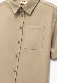 Camicia beige a maniche corte con bottoni, realizzata in tessuto morbido, con una piccola tasca sul petto e bottoni marroni, caratterizzata da un design con colletto classico.