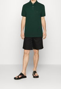 Tmavě zelené polo tričko se třemi knoflíky, černé plavky a černé pantofle nazouváky. Minimalistický design s pravidelným střihem a jemnou texturou.