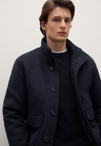 Marineblaue Fleecejacke mit hohem Kragen, Frontreißverschluss und Knöpfen. Verfügt über zwei Seitentaschen und eine strukturierte Oberfläche. Dünner Strickpullover darunter.