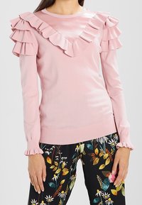 Un pull rose avec un col en V à volants et des manches à volants en cascade. Matière en tricot douce avec une coupe ajustée et des poignets côtelés.