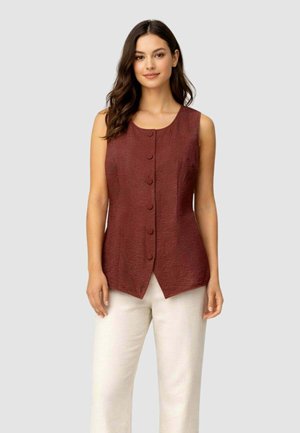 Donna con capelli lunghi e scuri che indossa una maglia senza maniche color melanzana con bottoni e pantaloni chiari, in piedi davanti a uno sfondo semplice.