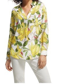 Chemisier blanc léger avec un motif floral composé de fleurs jaunes et roses, un col droit avec encoche et des manches longues, porté avec un pantalon blanc.
