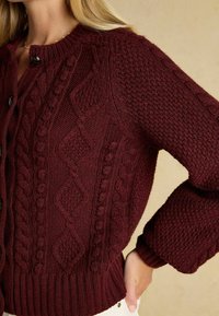 Bordová pletená cardiganka s textúrovaným káblovým vzorom, rebrovanými manžetami a okrúhlym výstrihom s uzáverom na gombík.