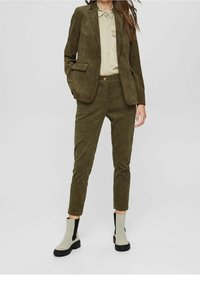 Zestaw marynarki i spodni z korduroju w zielonym oliwkowym kolorze, zestawiony z beżowymi botkami za kostkę z czarnymi akcentami. Pod spodem widać gładką koszulę z kołnierzykiem.
