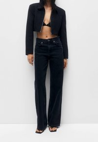 Woman wearing a cropped black jacket over a black bralette, high-waisted wide-leg black jeans, and black open-toe sandals standing against a white wall.