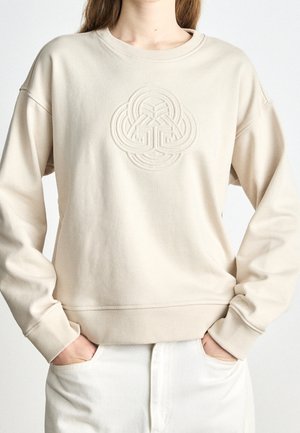 Personne portant un sweat-shirt beige avec un motif circulaire en relief et entrelacé sur la poitrine, associé à un pantalon blanc et les mains dans les poches.