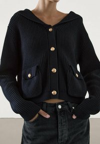 Kobieta w czarnym, grubym swetrze z guzikami w kolorze złota i kieszeniami z przodu, połączonym z wysokogatunkowymi czarnymi dżinsami, z rękami w kieszeniach.