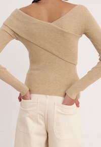 Beige väritetty neulepaita, jossa on olkapäät paljastava malli, pitkät hihat ja vartalonmyötäinen leikkaus. Yhdistetty valkoisiin korkeavyötäröisiin housuihin, joissa on takataskut.