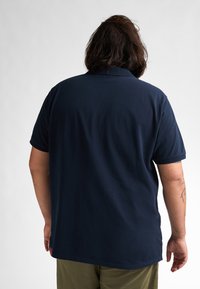 Navyblaue Poloshirt aus Baumwolle, mit Kragen, kurzen Ärmeln und einer lockeren Passform mit glatter Textur. Sichtbares Tattoo am Arm.