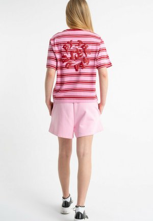 Osoba ubrana w różowo-czerwony pasiasty t-shirt z abstrakcyjnym czerwonym wzorem na plecach, różowe spodenki, białe skarpetki i czarne tenisówki, stojąca tyłem.