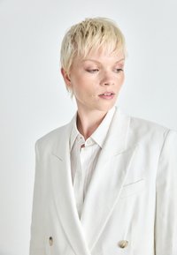 Blazer bianco doppiopetto con colletto a punta, dotato di bottoni dorati e una camicia color grigio chiaro a texture con colletto sottostante.