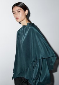 Teal silketop med et flydende, asymmetrisk design, der har en høj halsudskæring og draperede ærmer, suppleret af en glat, skinnende tekstur.