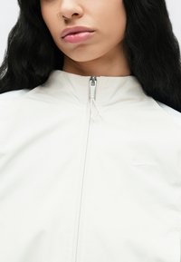 Λευκό τζάκετ με φερμουάρ κατασκευασμένο από ελαφρύ ύφασμ α με ψηλό γιακά, που διαθέτει ασημένιο φερμουάρ και διακριτικό λογότυπο Nike στο στήθος.