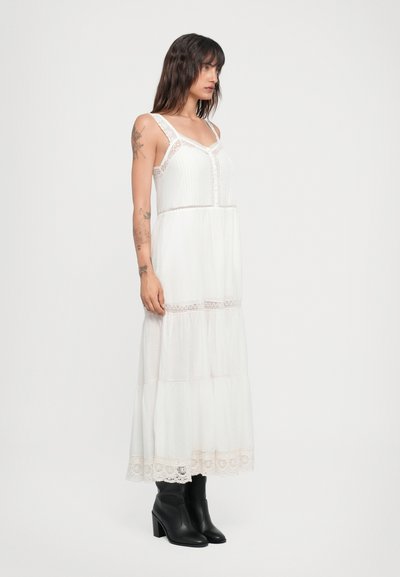 Vestido maxi blanco con detalles de encaje, diseño en capas y botones, combinado con botas de tacón negras. Tela suave, silueta sencilla.