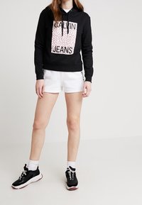 Sudadera con capucha negra con un gráfico de leopardo rosa y el texto "CALVIN JEANS". Combinada con pantalones cortos blancos y zapatillas negras con acentos blancos.