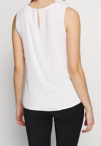 Blouse blanche sans manches avec un col rond et un détail en forme de clé à l'arrière, fabriquée dans un tissu léger au toucher doux.