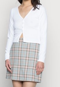 Bílý žebrovaný kardigan s dlouhým rukávem a výstřihem do V, se zápínáním na pět knoflíků, který je zkombinován s kostkovanou mini sukní v pastelových tónech modré, růžové a hnědé.