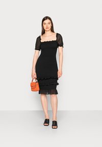 Vestido preto com mangas bufantes, textura franzida, bainha em camadas com babados, acompanhado de uma mala laranja e sandálias escorregonas pretas.