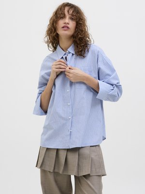 Chemise à rayures bleues et blanches avec une poche poitrine, assortie à un pantalon brun plissé. La chemise a une coupe décontractée et des manches retroussées.