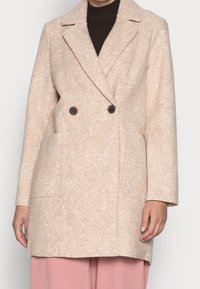 Béžový kabát z vlněné směsi v oversize stylu s klopovým límcem, dvouřadým zapínáním, dvěma velkými postranními kapsami a strukturovaným povrchem.