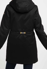 Svart lång kappa med huva, cinchade midja och detaljer i guldmetall. Slät textur och minimalistisk design.