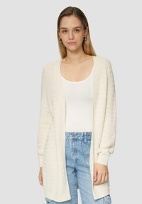 Gebreide, open voorkant cardigan in lichtcrème. Voorzien van ribbelstructuur, lange mouwen en een losse pasvorm. Gecombineerd met een wit topje en blauwe spijkerbroeken.