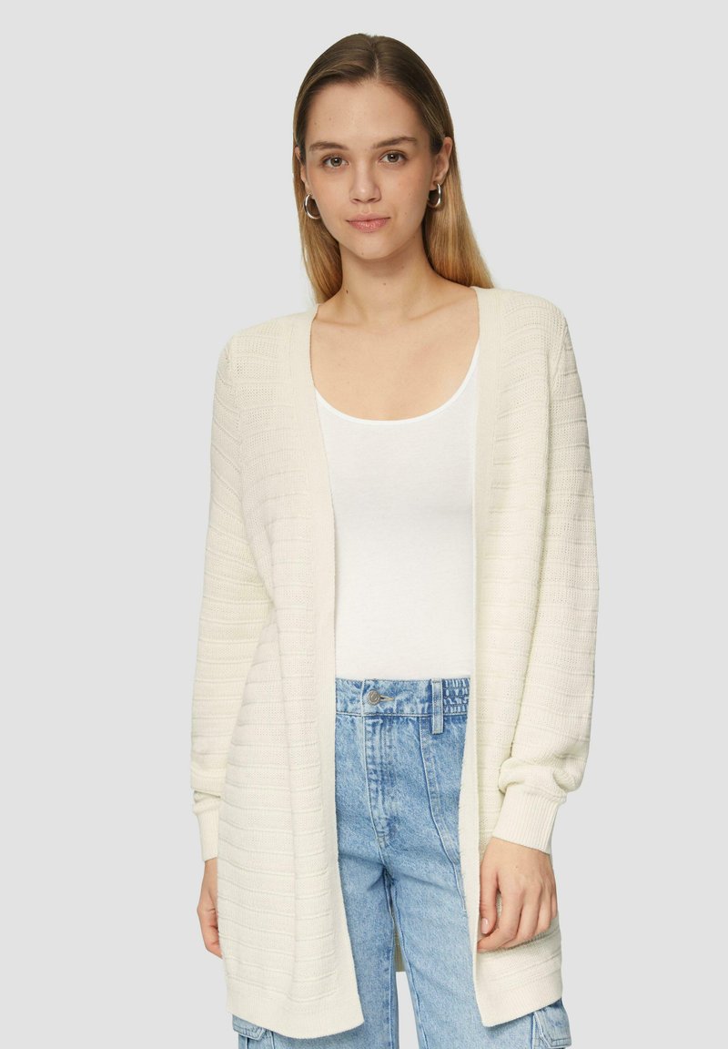 Gebreide, open voorkant cardigan in lichtcrème. Voorzien van ribbelstructuur, lange mouwen en een losse pasvorm. Gecombineerd met een wit topje en blauwe spijkerbroeken.