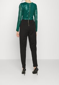 Haut à manches longues en sequins verts avec une fermeture éclair au dos, associé à un pantalon noir ajusté et des chaussures à talons noirs.