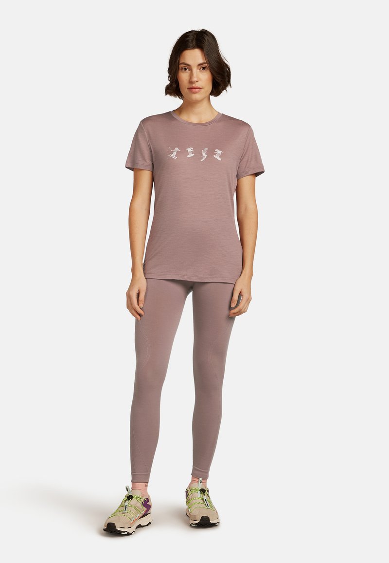 Kvinde stående iført en mauve kortærmet t-shirt med fire hvide skikoner, matchende leggings og beige trail-løbesko med grønne snørebånd.