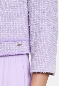 Lawendowa marynarka z tweedu o teksturowanym wykończeniu i skróconych rękawach. Wyrazisty złoty detal na dolnym brzegiem. Nałożona na pasującą spódnicę.