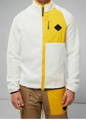 Mężczyzna w białej kurtce polarowej z żółtym panelem zapinanym na zamek i kieszenią na piersi, w parze z brązowymi spodniami z żółtą naszywką.