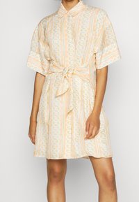 Šaty s krátkým rukávem a límečkem v pastelově oranžové barvě s bílými a zelenými vzory řetězů. Mají zavazování v pase a volný střih. Lehká texturovaná látka.